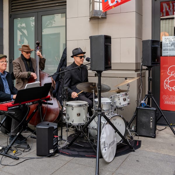Three musicians play jazz instruments, including a keyboard, upright bass, and drums, outside Cajun Claws.