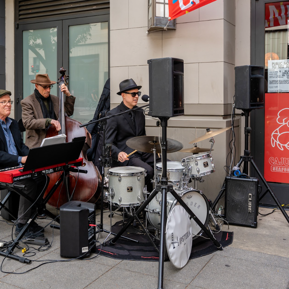 Three musicians play jazz instruments, including a keyboard, upright bass, and drums, outside Cajun Claws.