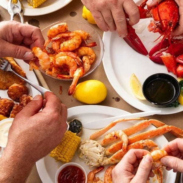Hands reaching for various seafood dishes, including lobster, crab, shrimp, and fried fish.