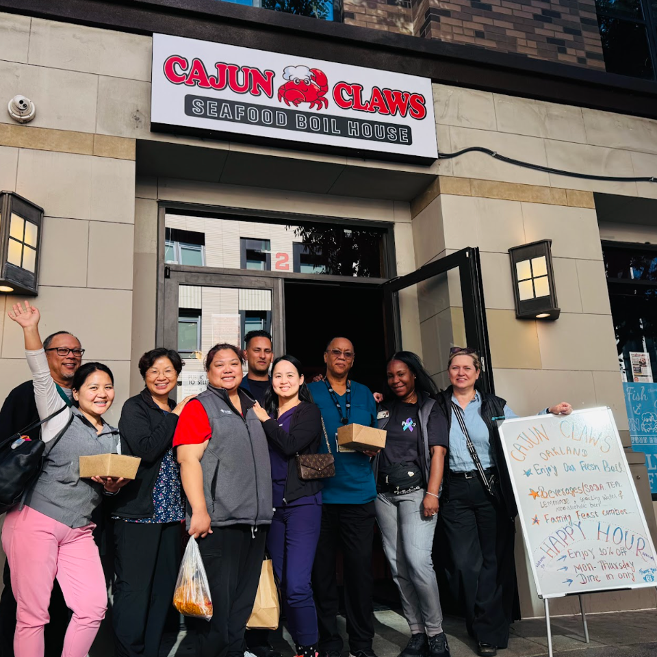 A group of diverse people smiling and holding food containers in front of Cajun Claws Seafood Boil House.