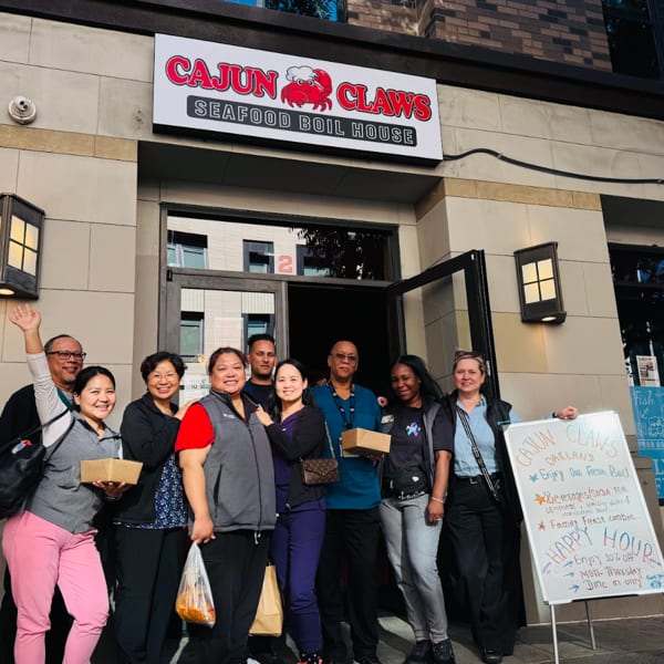 A group of diverse people smiling and holding food containers in front of Cajun Claws Seafood Boil House.