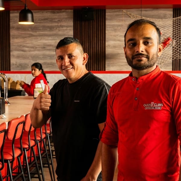 Two men smiling in a Cajun Claws restaurant, one giving a thumbs up at the bar.