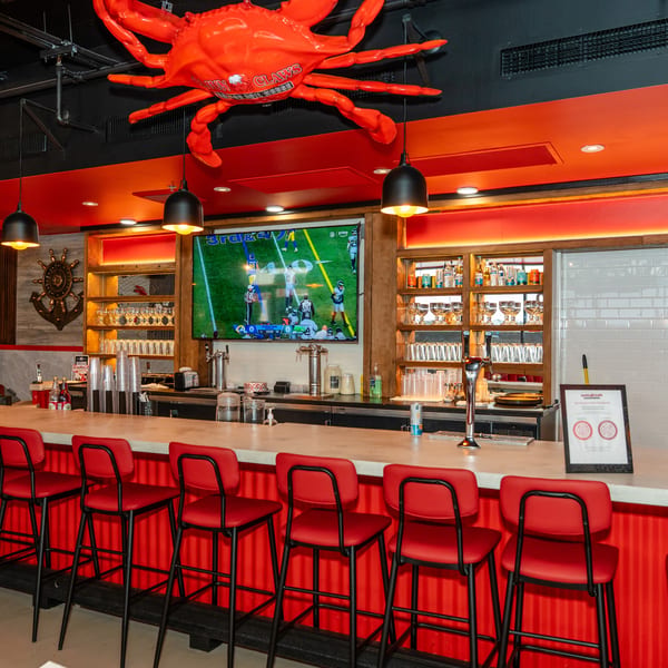 A vibrant bar with red stools, a large TV showing football, and a giant red crab decoration overhead.