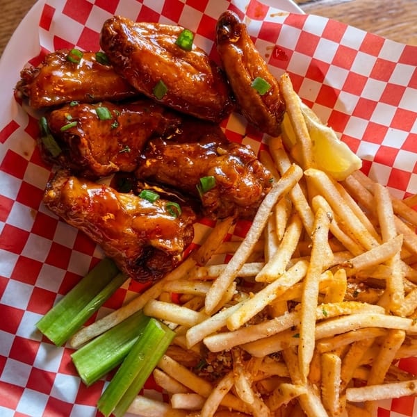 Sweet and sour chicken wings with french fries, celery sticks, and a lemon wedge on checkered paper.
