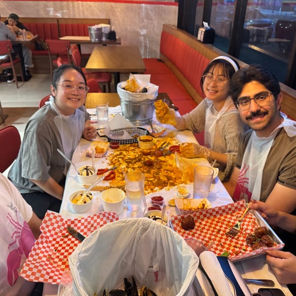 Five smiling friends wearing bibs and eating a messy seafood boil at a restaurant table.