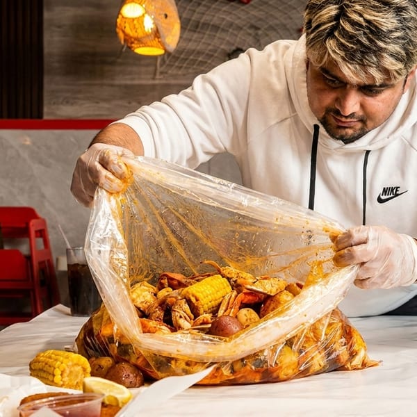 Man in gloves opening a large plastic bag filled with seafood boil, corn, and potatoes.